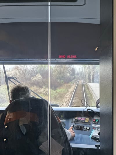 The image shows a view from inside the cabin of a train. Outside the window, the tracks extend into a landscape of trees and fields. A person, likely the train operator, is visible, wearing a dark jacket, and is seated in front of the train's control panel. The control panel features various buttons, displays, and switches. A timestamp of "31/03/25 08:02" is displayed in red on the left side of the window. The location cannot be verified as being in Gorna Mahala, Bulgaria, as no information in the photograph specifically identifies that geographic location.
