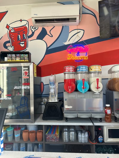 The image shows the interior of a small shop in Pozos, Costa Rica, located at the coordinates 9.951 latitude and -84.194 longitude. The shop appears to be a "cocktail and dreams" themed establishment, evident from the neon sign on the wall. The shop features a large, colorful mural depicting a cartoon glass of red liquid, with a smiling face, holding a bottle of red liquid in its hand. Behind the counter, there are three silver slushie machines, each with a glass bowl filled with different colored slushie mixtures. The bowls are labeled "Daiquiri de Fresa" (Strawberry Daiquiri), "Blue Ocean" (Blue Ocean), and "Piña Colada" (Pineapple Colada). Underneath the slushie machines, there is a counter with shelves stocked with various items, including plastic cups, straws, and bottles. The shop is decorated with a vibrant, lively atmosphere, suggesting it is a popular spot for locals and tourists alike.