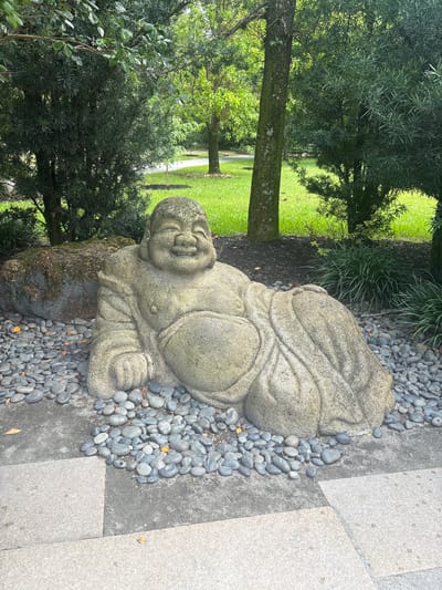 A stone statue of a smiling, reclining Buddha rests on a bed of small gray pebbles, partially surrounded by larger, light-colored paving stones. Behind the statue, a large, moss-covered rock sits near the base of a lush green tree. The background features a manicured lawn and more trees, with sunlight filtering through the leaves, suggesting an outdoor garden setting. This scene is located at the Morikami Museum and Japanese Gardens in Delray Beach, Florida, United States.