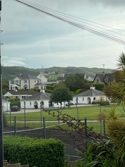This is a vertical, eye-level, outdoor, medium shot of a residential area in Dunfanaghy, Ireland. The scene depicts several houses with modern architecture, characterized by white exteriors, dark-tiled roofs, and large windows. Some houses feature arched doorways and manicured hedges. Cars are visible in the driveways and on the street.
In the foreground, a green sports field, likely a tennis court or similar facility, is enclosed by a dark fence. Lush green bushes and trees are abundant throughout the scene, adding to the natural beauty of the location. In the background, rolling green hills, characteristic of the Irish landscape, rise towards a cloudy sky. Overhead power lines are also visible in the upper left portion of the image.
The weather appears overcast, suggesting it is daytime but with limited direct sunlight. The overall impression is one of a peaceful, well-maintained suburban or semi-rural environment. No people are clearly visible in the frame, and there is no discernible text.