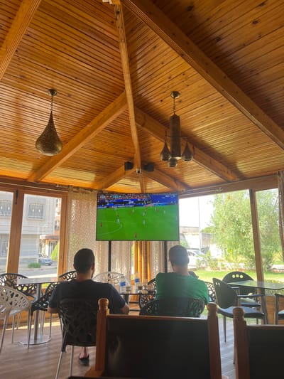 The image depicts the interior of a covered outdoor seating area, likely a cafe or restaurant, in El Hajeb, Morocco. The space is characterized by a prominent wooden vaulted ceiling with exposed beams and a warm, natural wood finish. Two men are seated, facing away from the camera and towards a large television screen displaying a soccer match. The man on the left is wearing a dark t-shirt and dark shorts, while the man on the right is wearing a green t-shirt and dark shorts. Both are seated in black, modern-looking chairs.
In the foreground, more seating and tables are visible. The TV is mounted on a stand and is centrally positioned in front of the seated patrons. The background shows large windows that look out onto a street and green trees, indicating an outdoor environment. The architecture of the building outside, with its light-colored facade and grid-like windows, suggests a common urban or suburban style. Two hanging light fixtures with an intricate, metallic design are suspended from the ceiling, adding to the aesthetic of the space. The overall atmosphere appears relaxed, with people engaged in watching the sports broadcast.