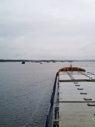 This image depicts a wide view of a body of water, likely a river or lake, with a fleet of barges and ships at anchor or moving slowly in the distance. The foreground is dominated by the deck of a large vessel, characterized by its grey, metallic surface, featuring numerous hatches and access points. The sky is overcast and grey, indicating a cloudy or possibly rainy day. The shoreline in the distance is lined with trees. The scene is set on the Svir River, near the city of Vytegra, Russia, a significant waterway in the region, part of the Vyshny Volochyok Water System. The presence of numerous vessels suggests a busy port or a staging area for cargo transport.
