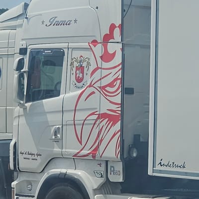 The image is a close-up, slightly angled shot of the side of a white semi-truck, taken outdoors in what appears to be a sunny environment. The truck has several decals and markings. On the upper portion of the cab, a stylized word "Inma" is visible in a metallic font, flanked by small stars. Below this, towards the front of the cab, a crest with a knight on a horse and a banner reading "AGÜERO" is displayed. A large, red graphic of a bird's head, possibly an eagle, is prominently featured on the side of the truck. Towards the rear of the cab, a smaller emblem is visible, and further back, a model designation "R660" is present. Below the "Inma" text, there is an inscription that appears to be "Angel M. Rodriguez Lujan" followed by "TARZANAS - ALMERIA". The rear trailer section of the truck is also white and has a logo or text in a cursive font that reads "indetruck". The background consists of a blurred white structure and a hint of greenery, suggesting an outdoor industrial or roadside setting. No people are visible in the frame.