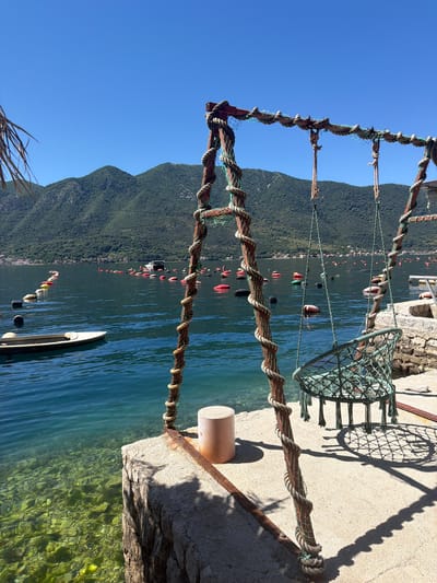 The image, taken in Dražin Vrt, Montenegro, shows a pier overlooking a body of water surrounded by mountains. A metal and rope structure stands on the pier, partially obstructing the view. A net-like chair rests on the pier to the right of the frame. Buoys are visible in the water in the background. The sky is clear and blue. The water is a turquoise color. A light-colored boat is visible in the lower center of the image.