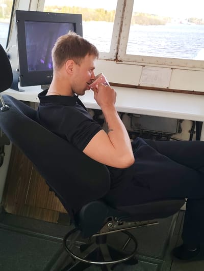 The image shows a man seated in a black chair, holding and drinking from a pink mug. He is wearing a black polo shirt and black pants. Beside him is a computer monitor. Through a window, a body of water and trees can be seen. Based on the location, the scene is likely inside a vessel on a waterway in Анхимово, Russia.