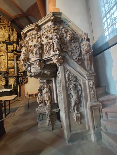 The image displays an intricately carved stone pulpit and its integrated staircase, positioned within a large religious edifice, specifically the St. Peter and Alexander Collegiate Church in Aschaffenburg, Germany.
The pulpit, constructed from light-colored stone, likely sandstone, features extensive Baroque-style sculptural decoration. Its multi-faceted body is adorned with numerous carved figures, depicting individuals in ecclesiastical or historical attire, religious scenes, angels, and cherubs, interspersed with floral and architectural motifs. The base of the pulpit is supported by standing carved figures.
An attached staircase, equally ornate, descends to the right, exhibiting further carvings of figures and decorative elements along its balustrade. The stairs themselves are plain, solid blocks.
In the background to the left, a large, multi-tiered altarpiece or reredos, also heavily carved with numerous figures and golden accents, is visible. The floor is covered with a checkerboard pattern of alternating light and dark square tiles. A plain wooden table with a white cloth and two yellow candles is visible near the altarpiece, cordoned off by a dark rope barrier.
The room features a high vaulted ceiling with visible structural elements in the upper left. A large rectangular window with a grid pattern is present on the right wall, admitting natural light and casting shadows on the wall and floor. No individuals are present in the scene.