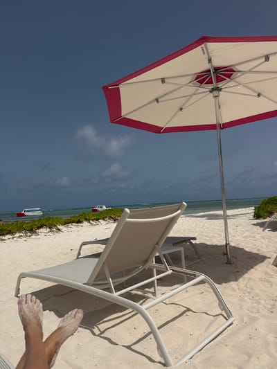 The image depicts a sunny day at a beach in Higüey, Dominican Republic. In the foreground, a pair of sandy feet are crossed, resting on a white lounge chair. The chair is situated on white sand, with shadows cast by an open beach umbrella, which has a white canopy and a pink trim. The umbrella is supported by a silver pole.
Further back, a line of green foliage runs along the edge of the beach. Beyond the foliage, the turquoise ocean stretches to the horizon under a partly cloudy, dark blue sky. Two white boats with red accents are visible on the water, one closer to shore than the other. The overall scene conveys a tranquil beach resort atmosphere.