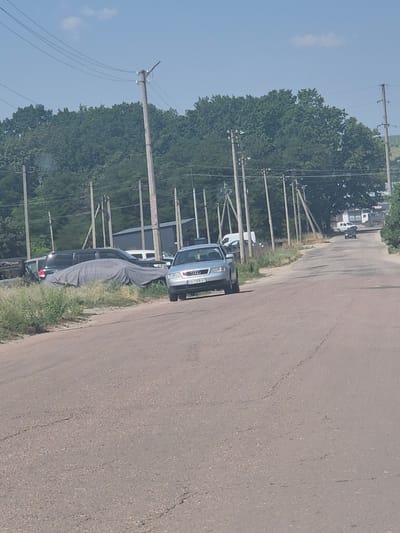 The image depicts a road in Pidhaitsi, Ukraine, with a line of utility poles and trees on the left side. A silver Audi sedan with a license plate reading "CA 7356 XT" is parked on the right side of the road, facing away from the camera. Several other vehicles are visible further down the road, including a white van and a dark-colored car. The road surface is asphalt with visible cracks and patches. The scene appears to be a rural or suburban area on a sunny day with a blue sky and scattered clouds.