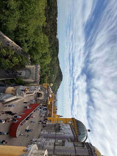 The image presents a high-angle, slightly tilted view of Pena Palace in Sintra, Portugal. A large, ornate castle complex dominates the foreground, featuring a mix of architectural styles and colors. A red carpet spans a courtyard, lined with numerous people. In the background, dense green forests cover the hills, extending towards a blue sky with scattered clouds. The sky takes up about half of the frame. The image appears to be taken from an elevated position within the castle itself.