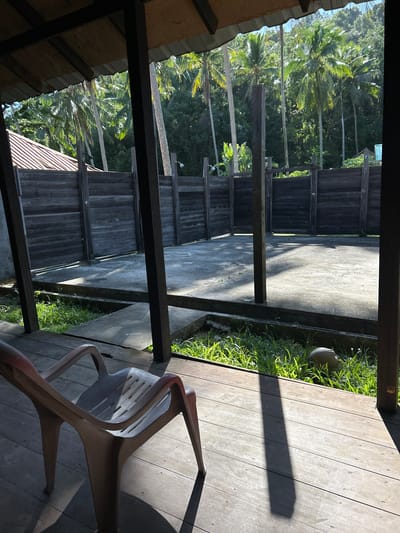 The image depicts an outdoor area under a covered structure, likely a shelter or veranda. The structure has a wooden deck in the foreground and wooden posts supporting a roof made of corrugated material and wood. In the middle ground, there is a concrete floor area surrounded by a dark wooden fence. Beyond the fence, lush green palm trees and other tropical vegetation are visible, suggesting a tropical or subtropical environment. The scene is illuminated by bright sunlight, casting strong shadows. In the foreground, a light-colored plastic chair sits on the wooden deck. The overall impression is of a rustic, open-air space in a tropical setting. The location is identified as Ban Khlong Hia, Thailand.