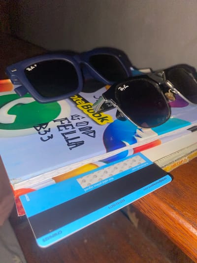 The image captures a collection of personal items arranged on a wooden surface, possibly a shelf or desk, in an indoor setting. The shot is taken at a tilted angle, looking down at the objects.
In the foreground, there are two pairs of sunglasses. The top pair is black with "Ray-Ban" visible on the right lens, while the bottom pair is blue, also featuring a "Ray-Ban" logo on its right lens. Behind the sunglasses is a light-colored notebook with "NoteBook" printed on its cover and a large green WhatsApp logo. Handwritten in black ink on the notebook are the words "GOOD FELLA B33".
To the right of the notebook, leaning against other unseen books or papers, is a blue and black financial card. Visible text on the card includes an email address "cards@moniepoint.com", a number "02016316270", "Authorised user", "Not valid unless signed", and multiple instances of the "Verve" logo. Another code, "CFMP0525", is partially visible on the bottom right edge of the card.
No activity is depicted, and no people are present. The lighting suggests an indoor environment. Given the presence of "moniepoint.com" and "Verve" branding, which are associated with Nigerian financial services, the items strongly indicate a location context in Nigeria, specifically Jama A as provided.