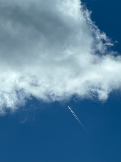 A wide shot captures a vast expanse of deep blue sky with a large, white, fluffy cloud formation dominating the upper half of the frame. Below and to the right of the cloud, a white contrail stretches diagonally from the lower left to the upper right, indicating the path of an airplane. The image does not contain any information to place it in Gela, Bulgaria, or any other specific urban location.