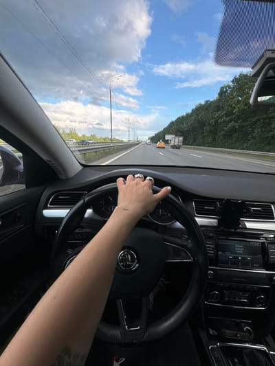 A first-person perspective from inside a car shows a hand with rings on the steering wheel. The driver's left arm, with the word "now" tattooed on the forearm, is visible as the car moves along a highway. The car's dashboard displays a radio with the song "Supergirl" by Reamonn playing. Outside, the sky is partly cloudy, and trees line the highway. The scene is identified as being in Sapronovo, Russia.