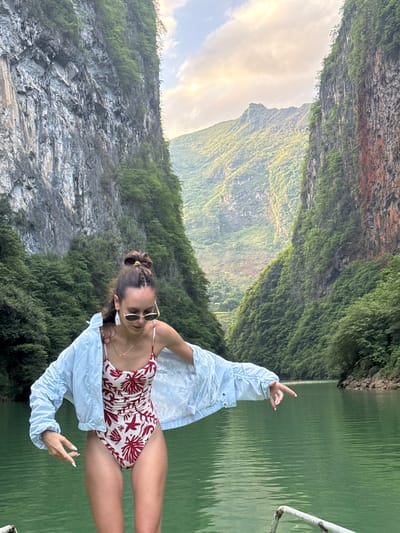 A woman is standing on a boat on a river in the Đồng Văn Commune, Vietnam. She is wearing a light-colored floral one-piece swimsuit and a blue jacket, with sunglasses on her face. She has a bun and a necklace. She is standing on a boat on the river, gesturing with both hands towards the mountain landscape. The environment includes lush greenery covering steep rock formations. The sky is partially covered by clouds. The river has a green color.
