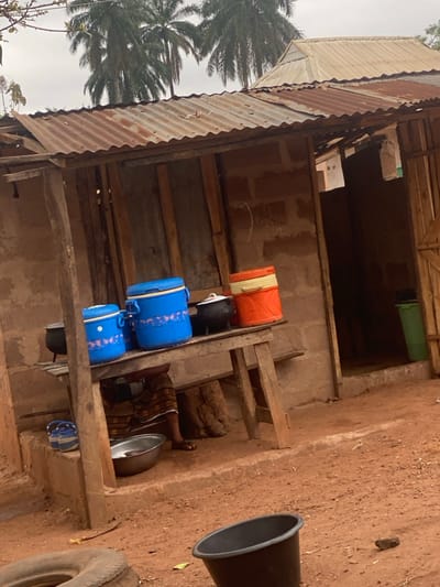 A low-angle shot displays a dilapidated structure with a corrugated iron roof. Inside, storage containers in various colors and sizes are stacked on wooden shelves. A person lies seemingly asleep amidst the containers and a metal bowl. Outside, bare reddish-brown earth is visible, along with a black bucket and a tire. A palm tree is visible on the left. The scene conveys a sense of poverty and simple living.