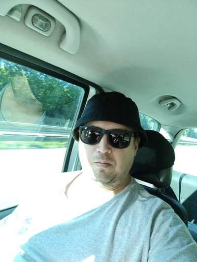 A man wearing a black bucket hat and sunglasses takes a selfie inside a car, appearing to be on a highway. The sun is shining brightly, casting shadows inside the vehicle. The man's reflection is visible in the car window, showing trees and other vehicles passing by. The interior of the car shows a light-colored headliner with a grab handle and a light fixture. The man is wearing a gray t-shirt. Based on the context provided, the location is the city of Psáře, Czechia, likely on a road that runs through or near it.