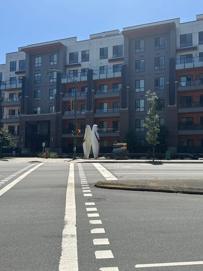 The image depicts a street scene in front of a modern apartment building in Port Moody, Canada. The building features multiple stories with a combination of grey and white exterior panels, accented by orange balconies and dark vertical columns. Two abstract sculptures, resembling elongated teardrop shapes, stand prominently on the sidewalk near the building's entrance. A traffic light with a pedestrian signal displays a red walking figure. The street in the foreground has clear lane markings, including solid white lines and dashed white lines. A few trees and some landscaping are visible on the property. The sky is clear and blue, indicating a sunny day.