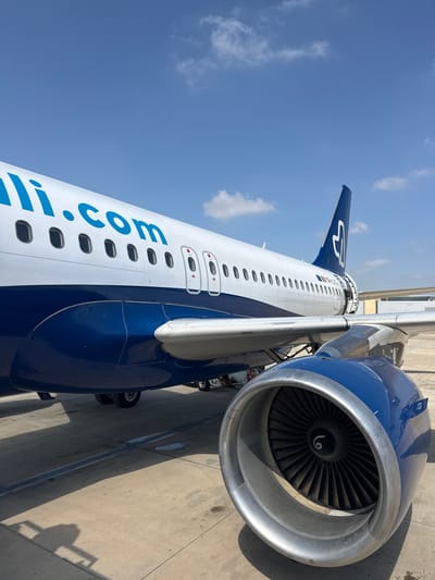 The image shows an aircraft, likely at Sdot Dan Regional Council, Israel. The blue and white fuselage is prominently featured, with the website address "lill.com" visible along the side. An engine is visible on the right. The tail of the aircraft, also blue and white, is visible in the background. The aircraft is parked on a concrete surface. The sky is blue with scattered clouds.