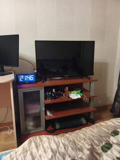 The image shows a bedside table with a TV on top. There are several items on the shelves of the table, including a digital clock that displays "21:46", a glass with a clear liquid, a bottle of cologne, and a package of wet wipes. There is also a black bag on the bottom shelf. To the left of the table is a computer monitor. The room appears to be a bedroom, with a bed covered in a white comforter visible in the foreground. The walls are painted a light color. The lighting in the room is dim. The image appears to have been taken in the city of Kommunar, Russia.