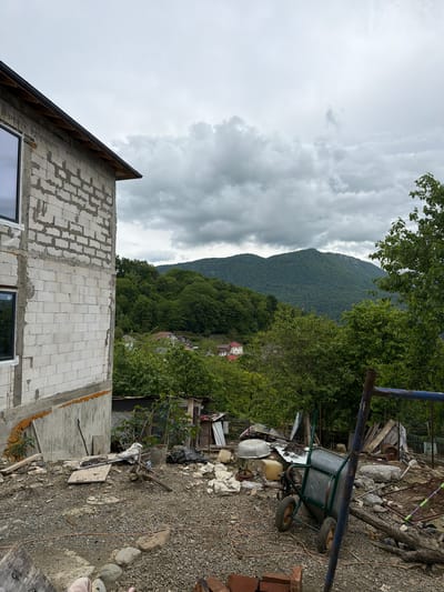 The image depicts an overcast scene in what appears to be a rural or semi-rural area. In the foreground, there is an unfinished building under construction. The walls are made of white concrete blocks, and window frames are visible. In the midground, there's a yard area filled with gravel, construction debris, and miscellaneous objects, including a wheelbarrow. A lush, green forested area is visible beyond the yard, with a mountain in the background. Based on the prompt, this location is identified as being in Galitsyno, Russia.