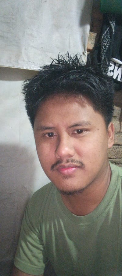 This is a portrait of a young man indoors, possibly in Kemuning, Indonesia. He is looking directly at the camera with a neutral expression. He has dark, spiky hair and a mustache and a small patch of stubble on his chin. He is wearing a green t-shirt. The background is a textured, light-colored wall with some discoloration and a dark object with a patterned fabric in the upper right corner. The lighting is somewhat dim, casting shadows on the man's face and the wall.