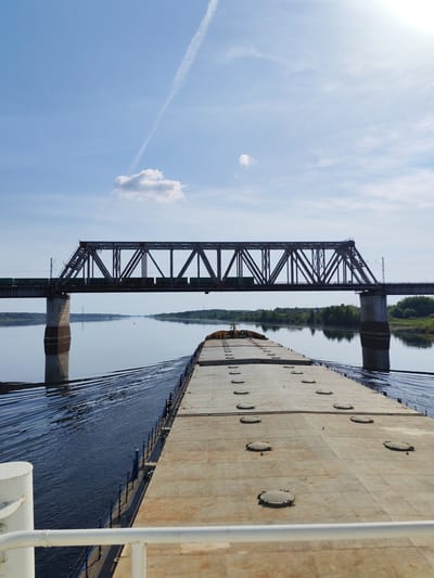 The image depicts a large, flat-topped barge navigating a wide river in Tarkanovo, Russia. The barge is heading directly toward a dark, steel truss bridge, which appears to carry rail traffic as evidenced by train cars visible on the bridge. The water is relatively calm, reflecting the bridge and sky. The sky is light blue with a few scattered clouds and a contrail. Vegetation is visible along the riverbanks. The view is captured from the front of the barge. The barge's deck is a uniform light brown/beige color with a series of circular manhole covers. A white railing is visible in the foreground on the lower left side of the frame.