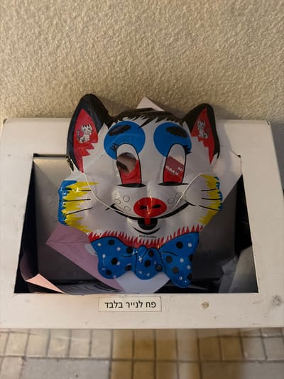 A multi-colored plastic mask, resembling a cat with clown-like features, is centrally positioned within a white, open-top container, identified as a paper recycling bin in Harish, Israel. The mask displays a white base with black ears, blue circular eyes, a red nose, and a red mouth. Small white cartoon cats are depicted on the red inner sections of the ears. A blue and black polka-dotted bowtie is visible below the mouth, and yellow and blue wavy lines adorn the cheeks. The text "MADE IN CHINA" is present on the mask's chin. Behind the mask, a pink piece of paper with a simple leaf design is partially visible. A white label affixed to the front of the container displays the Hebrew text "פח לנייר בלבד," translating to "Paper bin only." The container rests on a light-colored tiled floor, with a textured, light-colored wall forming the background.