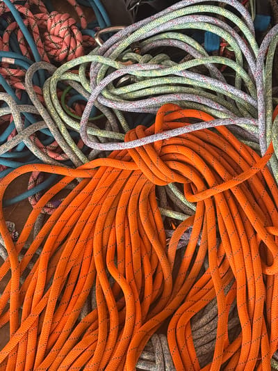 The scene, located in the city of Tayshet, Russia, presents a dense, overhead view of numerous tangled climbing ropes. A prominent, bright orange rope with a black speckled pattern dominates the foreground and midground. Intertwined within this mass are several other ropes of varying colors, including light green, light blue, pink, gray, and purple, all featuring a braided or woven sheath texture with distinct flecking patterns. A silver-colored metallic carabiner is partially visible amongst the lighter colored ropes in the upper left quadrant. The ropes are irregularly coiled and intermingled, resting on an indistinct dark surface. No individuals or specific environmental markers beyond the ropes are discernible.