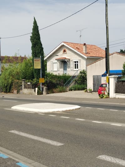 A long, thin, dark green evergreen tree stands tall on the left side of the image. In front of the tree, a yellow sign displays the words "ANTIQUITES BROCANTE" along with other text in French. To the right of the tree, a light-colored house with a red tiled roof is visible. The house has white trim around the roofline and shutters on some windows. A set of stairs leads up to the front entrance. A small red car is parked in the driveway of an attached structure to the right of the house. A blue awning is visible above a shop window to the right of the red car. The scene appears to be a street in a town or village, possibly in Le Passage, France, as indicated by the text on the sign. The road in the foreground has white lane markings and a white raised curb. The sky is overcast.