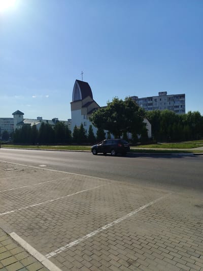 The image depicts a street scene in the city of Salihorsk, Belarus, on a clear, sunny day. In the foreground, a paved parking lot with white painted lines is visible. To the right of the parking lot is an asphalt road where a black SUV is driving. Behind the road, a row of trees lines a green strip of land. Further in the background, a modern church with a distinctive arched roof and a cross on top stands prominently. Adjacent to the church are several apartment buildings. The sky is a vibrant blue with a few wisps of clouds. The overall impression is one of urban development in a suburban setting under good weather conditions.