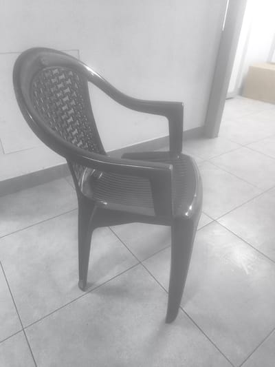 The image shows a single black plastic chair positioned in a room with tiled flooring. The chair has a woven pattern on its backrest and a solid seat. Its arms curve upwards and outwards. The chair is the primary object in focus, placed in the center of the frame.
The setting appears to be indoors, possibly an office or a waiting area, given the tiled floor and the white wall in the background. There is a hint of a doorway or opening on the right side of the image, with a light-colored object, potentially a cardboard box, visible beyond it. The lighting is somewhat diffused, with bright areas and shadows suggesting it might be daytime, though no windows are directly visible.
There is no activity or event taking place; the scene is static and still. No people are present in the image.
Notable details include the texture of the chair's backrest, the sheen of the plastic material, and the grid pattern of the floor tiles. The overall mood is neutral and utilitarian, with no strong emotional cues.
No visible text is present in the image. The location context provided is Khatay Raion, Azerbaijan, but there are no specific visual cues in the image itself to confirm this location.