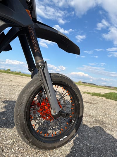 The image depicts the front wheel and suspension of a black and orange motorcycle on a concrete surface, possibly a road or parking area. The sky is visible in the background, exhibiting scattered clouds and blue patches. The foreground shows details of the motorcycle's front suspension, including the "BOLD" branded fork tubes. The front wheel has orange spoke nipples and a "VMAX" branded tire. The surrounding environment appears to be a rural or semi-rural area, with a grassy or agricultural field visible on the horizon. There is no indication of Voysil, Bulgaria or any human presence.