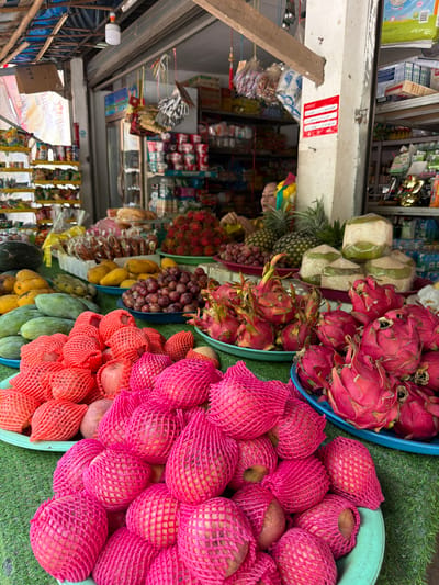 A fruit stall is set up in front of a store in Xieng Men cultural village, Laos. In the foreground, various fruits are displayed on green and blue trays. A large pile of red-netted apples is on the left, with more pink-netted apples and what appear to be peaches or nectarines in the center and right. Behind these, a large quantity of dragon fruit is visible on a blue tray, with more in the background. To the left of the dragon fruit are grapes on a blue plate and mangoes on a yellow plate. Further back, several green mangoes and bananas are displayed. A watermelon is visible on the far left. Behind the fruit display, the interior of a store is visible, stocked with shelves of packaged goods. A woman is partially visible behind a counter in the back, attending to a customer. The stall appears to be under a thatched roof supported by wooden beams. The overall scene depicts a vibrant marketplace filled with fresh produce.