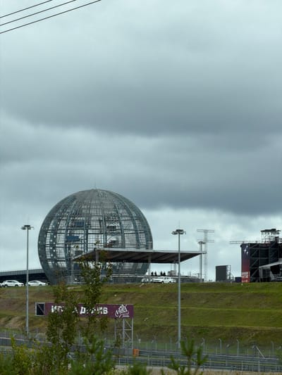 The image shows an outdoor scene under a cloudy sky, likely in Novozhilovo, Russia. In the background, a large, spherical, wireframe structure dominates the view. To the right of this sphere, a stage structure with lighting rigs and large screens is visible. In the mid-ground, a tiered grassy hill slopes upwards. Several white vehicles are parked on the crest of the hill, near a covered platform. People are standing on this platform. A sign with white text on a purple background is also present on the hill. In the foreground, there is a glimpse of a race track with fencing. Vegetation, including trees and grass, is scattered throughout the scene. Three dark, parallel lines are visible in the upper left corner of the image, possibly power lines.