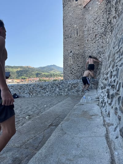 In Collioure, France, two shirtless men, one in black shorts with white stripes and another in black shorts, are climbing a stone wall near a tower. The man in the striped shorts is in the foreground, ascending a step. The other man is further up, entering an arched opening in the wall. Two plastic water bottles are visible on the steps below them. The background shows a view of the town with buildings and hills.