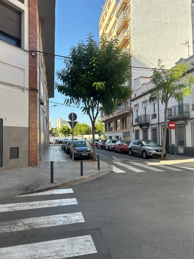 The image depicts a street scene in Castelló de la Plana, Spain. On the left, a building with a brick facade and a grey lower section is visible, with a rolling security shutter on a window. The sidewalk has a textured paving pattern and is lined with bollards. A pedestrian crossing with white stripes is in the foreground.
To the right of the sidewalk, several cars are parked along the street, including a black hatchback with license plate "0753 FVB," a red sedan, a white sedan, and a silver SUV. A large tree with green foliage stands in the middle of the scene. Beyond the parked cars, a multi-story building with balconies and cream-colored walls is prominent. Another building with a white facade and dark trim is to the far right, featuring balconies with wrought-iron railings and a red and white circular "no entry" sign. Trees and other buildings, some with balconies and awnings, are visible in the background, along with a clear blue sky. The overall impression is an urban street scene during daylight, with a mix of residential and commercial architecture.