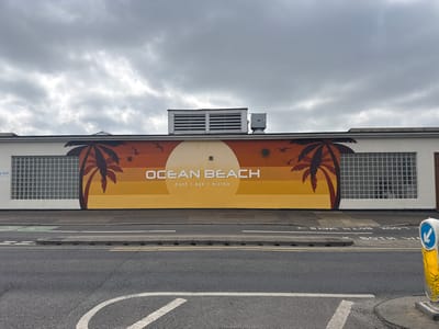 An outdoor, eye-level shot on an overcast day in Southend-on-Sea, United Kingdom, captures a low-rise commercial building adorned with a vibrant mural, bordering a multi-lane road.

The building's facade is predominantly white, featuring large sections of glass block windows on either side of the central mural. A prominent mural covers the middle portion of the wall, depicting a stylized beach scene. Against a backdrop of warm, horizontal gradient stripes transitioning from a burnt orange at the top to a golden yellow at the bottom, a large off-white circle represents the sun. Silhouettes of two palm trees, one on the left and one on the right, frame the scene, with three small bird silhouettes flying near the top left of the sun. The words "OCEAN BEACH" are centrally placed in large white capital letters within the sun motif. Below it, in smaller white capital letters, reads "CAFÉ | BAR | BISTRO". Above the left-hand glass block window, a small blue sign with white text indicates "TOILETS". On the roof, several industrial-looking ventilation units are visible against the cloudy sky.

In front of the building, a wide asphalt road stretches across the foreground. It features clear road markings, including solid white lines, dashed white lines, and a large white turning arrow. Part of the words "LOOK BOTH WAYS" are painted in white on the road, though partially obscured and appearing inverted in the main image. A grey concrete kerb separates the road from a narrow green-painted cycle path and a grey pavement area. On the far right, a yellow and black bollard-like structure with a blue circular sign featuring a white downward-pointing arrow stands at the edge of the road, indicating a traffic direction or hazard. No people or vehicles are visible, suggesting a quiet moment.
