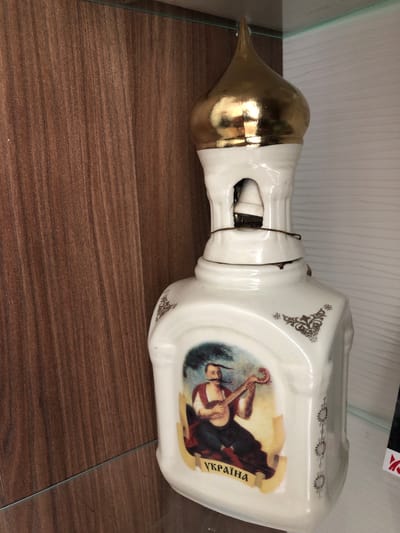 The image shows a close-up of a white ceramic bottle shaped like a church dome with gold accents. The bottle features a painting of a man in traditional clothing playing a lute and the word "УКРАЇНА" (Ukraine) written in yellow. The bottle is placed on a glass shelf, with a wooden surface visible to the left and a white striped surface to the right. There is no indication of the city of Leszno, Poland, in the image.