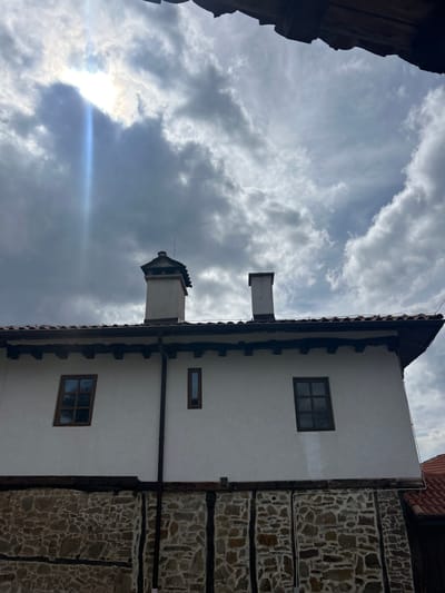 The image displays a two-story building in Gigintsi, Bulgaria, viewed from a low angle against an overcast sky. The lower section of the building features irregular stone masonry with dark, vertical, and horizontal timber beams. The upper section consists of a white plastered wall. Two rectangular windows with dark frames are present on the upper wall: a larger, multi-pane window on the left and a narrower, single-pane window on the right. A dark vertical drainpipe runs down the right side of the white wall, extending to the stone base. The building is topped with a terracotta-tiled roof, featuring dark-colored eaves with decorative supports. Two chimneys rise from the roof: a larger, ornate chimney with a finial/lightning rod on the left, and a simpler, rectangular chimney on the right. A small portion of an adjacent red-tiled roof is visible on the far right. The sky is largely covered by grey and white clouds, with the sun partially visible in the upper left, producing a distinct lens flare with a blue streak. A dark, wooden structure overhangs the top edge of the frame, partially obscuring the sky. No individuals or active interactions are visible within the frame.