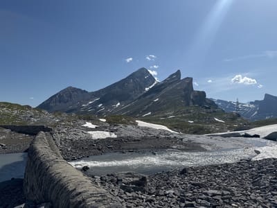 The image depicts a mountainous landscape in Leukerbad, Switzerland, under a clear blue sky with sparse clouds. In the foreground, a rocky embankment lines a flowing body of water. A low, curved stone structure, possibly a dam or retaining wall, is prominent in the lower left quadrant. The terrain is characterized by dark grey rocks, patches of snow, and sparse green vegetation. In the mid-ground, the water appears somewhat turbulent. Further back, imposing, jagged mountain peaks dominate the horizon, with remnants of snow visible on their slopes. A few distant structures and a transmission tower are also visible on the slopes. The overall scene conveys a sense of rugged natural beauty and a high-altitude environment.