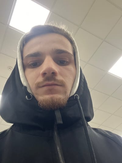 A man wearing a black hooded jacket is looking directly down at the camera. The hood is up, revealing a white lining. He has light brown hair, a light complexion, and a short beard. The background consists of a brightly lit, tiled ceiling, suggesting an indoor location.