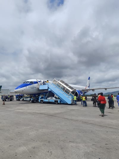 An overhead shot shows a passenger airplane parked on a tarmac at an airport in Gwaska, Nigeria. A set of blue mobile stairs is attached to the plane's door, and several passengers are seen walking up the stairs. Some airport personnel in high-visibility vests are directing traffic and assisting passengers. Luggage carts are also visible on the tarmac near the aircraft, indicating that baggage is being loaded or unloaded. The sky is overcast with dramatic clouds.
