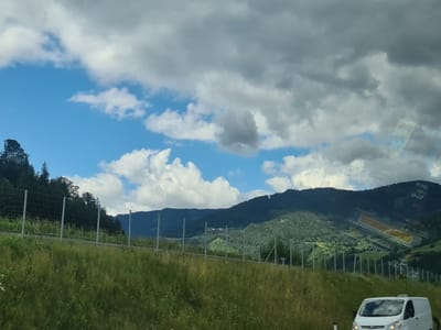 The image captures a scenic view of a mountainous landscape in St. Georgen ob Judenburg, Austria. In the foreground, tall green grass lines the side of a road, with a white van visible in the lower right corner. A wire fence runs horizontally across the mid-ground, separating the grassy area from a densely forested hillside. The forest extends up the slopes of rolling, dark green mountains, creating a textured backdrop. The sky above is a mix of bright blue with scattered white and gray clouds, suggesting a partially cloudy day. The overall scene conveys a sense of natural beauty and rural tranquility.