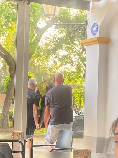 The image, captured on June 24, 2024, in San Pedro De Alcántara, Spain, depicts a scene through a large window or glass partition, likely from a cafe or restaurant. In the foreground, two men are visible from the back. The man on the left has grey hair and wears a black t-shirt with some text that is partially obscured. The man on the right has a shaved head and is wearing a dark t-shirt and light-colored shorts. They appear to be standing close together, with their arms touching or hands possibly clasped.

Behind them, the outdoor environment is lush with green trees and foliage, bathed in bright sunlight, suggesting a warm, possibly late afternoon or early morning setting. Sunlight filters through the leaves, creating a dappled effect. A black car is partially visible in the background.

Inside the establishment, on a table in the foreground to the left, a small wooden box with a patterned design sits on a dark-colored table, which is supported by black metal legs. To the right, a white pillar with decorative moldings is prominent, and an ADT security sign is affixed to it. The interior space is minimally visible, with hints of seating and structural elements. A portion of a person's face, looking towards the camera, is visible in the far right foreground, suggesting another patron or person within the establishment.