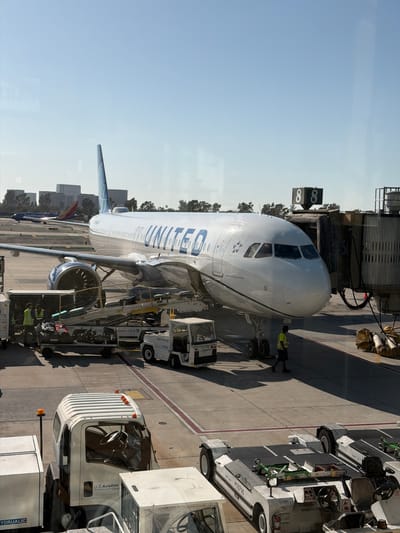 A United Airlines Airbus A320 aircraft is parked at a gate at John Wayne Airport (SNA) in Santa Ana, California, USA. The plane is connected to a jet bridge, and airport personnel are visible around it. Luggage carts, a tug vehicle, and other airport equipment are on the tarmac. In the background, other aircraft and airport buildings are visible. The sky is clear and blue.