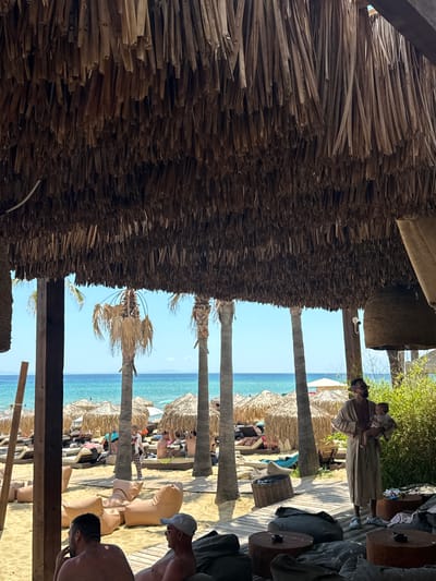 The image shows a beach scene at a resort in Nea Peramos, Greece. In the foreground, two men are seated on beanbag chairs under a thatched roof. To the right, a man wearing a robe is holding a baby. In the middle ground, there are beach chairs and umbrellas set up on the sand, with people relaxing. Palm trees and the turquoise waters of the Aegean Sea are visible in the background. The overall atmosphere is relaxed and summery, suggesting a vacation setting.