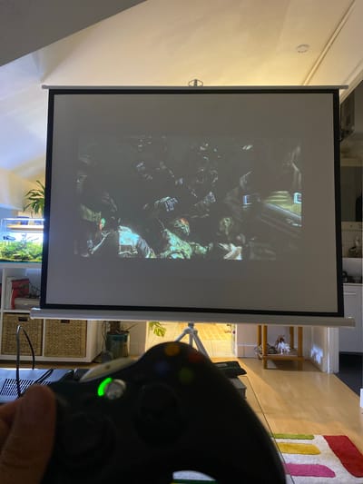The scene depicts an interior residential space in Boulogne-Billancourt, France, focused on home entertainment in dim lighting conditions. In the immediate foreground, a person's right hand holds a black Xbox 360 controller; the controller's guide button emits a prominent green glow, indicating it is active. A black cable extends from the controller area towards the bottom left.
Dominating the central midground is a large, white pull-down projector screen with a black border, displaying a dark, complex scene. The screen content appears to be from a video game or film, showing multiple figures in tactical or military-style gear, some with helmets and what look like goggles, positioned amidst a dimly lit environment with muted greens, browns, and grays. The screen is supported by a visible light-colored tripod stand at its base.
The background reveals elements of a furnished room. To the left of the projector screen, a white shelving unit holds a large, brightly lit planted aquarium on its top shelf, containing aquatic plants and a dark-colored fish. Below the aquarium, two woven wicker baskets are visible within the shelving unit, along with books and other items. A large green potted plant is situated behind the aquarium. To the right of the screen, a light wooden side table with two tiers displays a white animal figurine, possibly a unicorn or horse, and a small potted plant. Beyond this, a white doorframe is partially visible. The room features light wooden flooring, with a small rectangular rug in the bottom right corner displaying horizontal stripes in red, pink, yellow, and green. The ceiling is white, and warm ambient lighting illuminates the space.