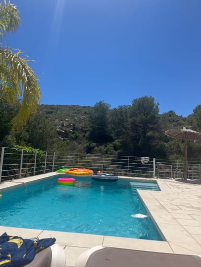 The image depicts a rectangular swimming pool in a residential setting, likely in el Poble Nou de Benitatxell/Benitachell, Spain, given the local flora and architectural styles commonly found in the area. The pool is filled with clear, turquoise water, containing several inflatable pool toys including a black and white tire, a pineapple and a multi-colored ring. The pool is surrounded by light-colored tiles and a white metal fence that separates the pool area from the natural landscape. The background reveals a lush green hillside with trees and rocky outcroppings, typical of the Mediterranean region. Above the hill, a clear blue sky dominates the upper left portion of the image. A straw sun umbrella and a metal chair are seen in the background, suggesting a leisure or vacation environment. In the foreground, part of a lounge chair and a blue shirt with yellow motifs on it can be seen.