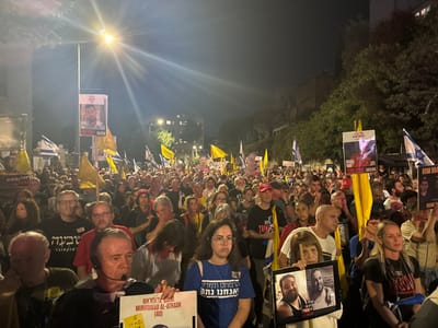 The image depicts a large, dense crowd gathered at night on an urban street in West Jerusalem, Israel. Hundreds of individuals of diverse ages and genders are visible, participating in what appears to be a vigil or protest for missing persons and hostages.

The scene is illuminated by prominent overhead streetlights, casting distinct light and shadows. Participants are tightly packed across the width of the street, extending into the background where urban buildings and trees are discernible.

Numerous objects are held by the crowd:
*   Many Israeli national flags (blue and white) are interspersed throughout the gathering.
*   A significant number of plain yellow flags, often associated with calls for the return of hostages, are also visible.
*   Attendees hold various signs and banners. These signs prominently feature photographic portraits of individuals, often accompanied by names (e.g., "MUHAMMAD AL-ATRASH (40)" is clearly visible on one sign held by a man in the foreground; another banner shows "ROM BR..." with a photo and "BRING THEM HOME NOW"). Text on signs is presented in both Hebrew and English, consistently conveying messages such as "BRING THEM HOME NOW."

People in the foreground exhibit somber and focused expressions, indicative of the serious nature of the assembly. There are no overt interactions or actions beyond standing and holding signs/flags, suggesting a peaceful, organized demonstration.