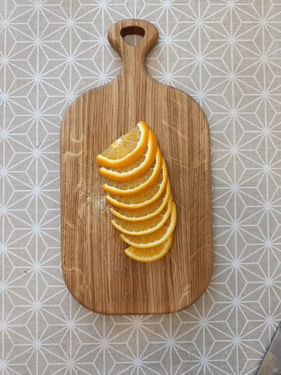 This media file shows a top-down view of a wooden cutting board on a textured, light-colored tablecloth with a geometric pattern of white lines forming star-like shapes. Placed on the cutting board are several thin slices of orange, arranged in a fanned-out pattern. The orange slices are vibrant in color, with visible pulp and rind. A small amount of what appears to be sugar or salt crystals is scattered on the cutting board near the orange slices. The cutting board has a handle with a hole and exhibits the natural grain of the wood. The setting appears to be indoors, likely a kitchen or dining area. No people are visible. The lighting suggests it is daytime, with no indication of specific weather conditions. There is no visible text in the image.